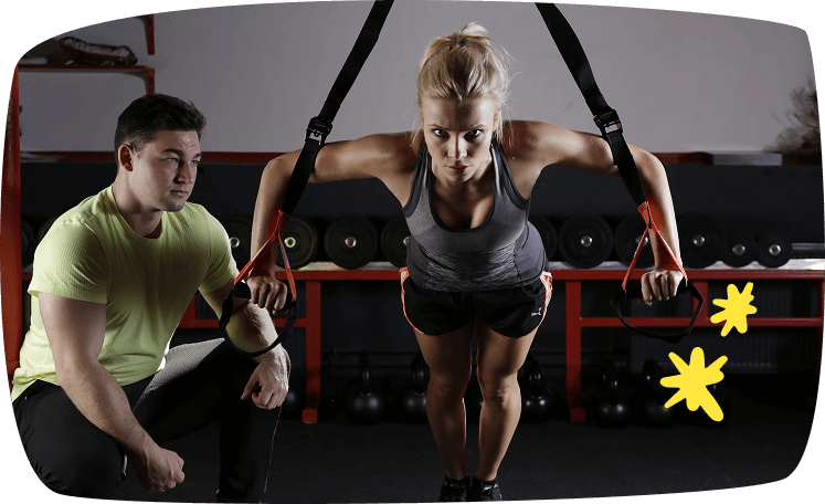 Entrenadora personal guiando a una mujer en un ejercicio de TRX en el gimnasio, ejemplo de trabajo extra como coach deportivo.