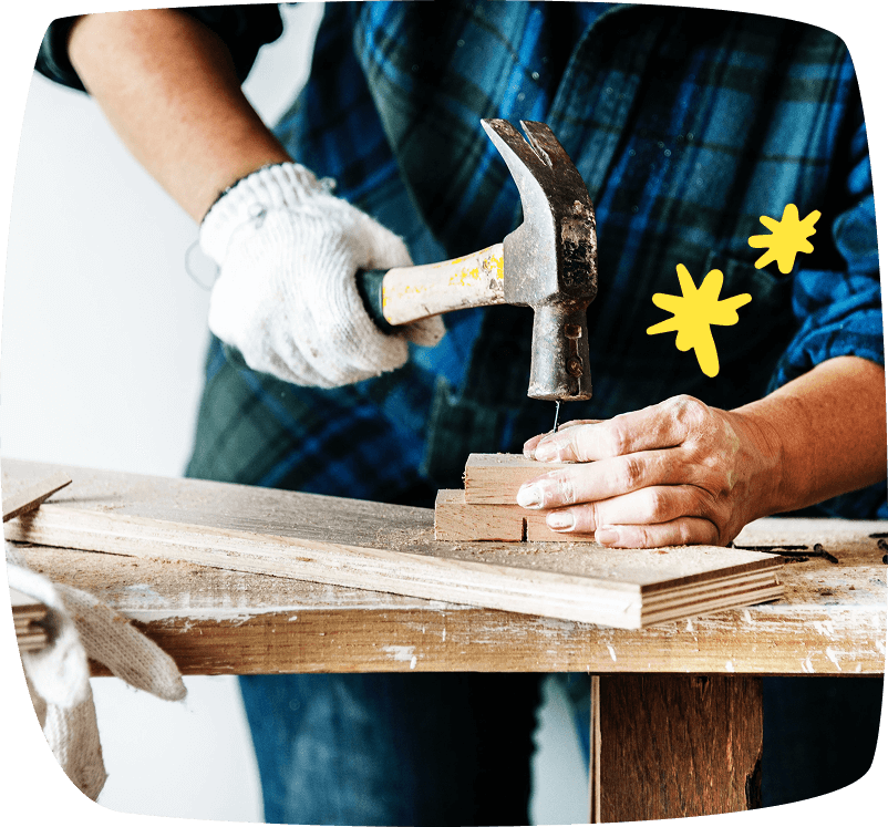 Persona clavando un clavo en un bloque de madera sobre un banco de trabajo, representando trabajos de manitas y bricolaje.