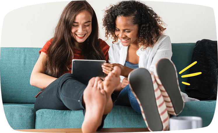 Dos amigas sentadas en un sofá riendo mientras miran una tablet, compartiendo un momento relajado.
