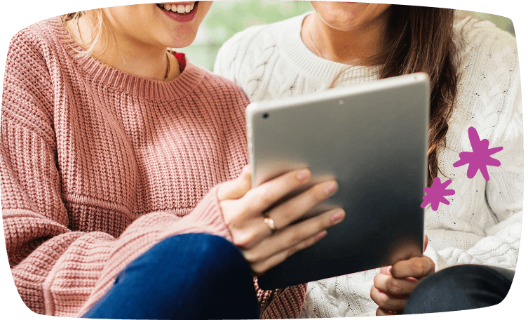 Dos amigas sentadas una junto a la otra mirando una tablet con sonrisas, compartiendo contenido digital.