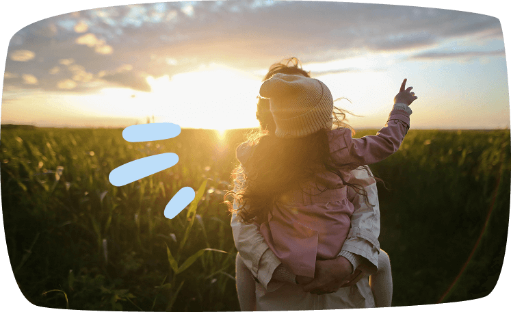 Madre con su hija en brazos señalando el atardecer en un campo abierto, momento familiar y natural.