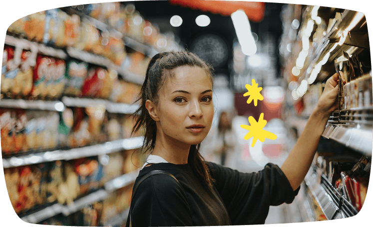 Estudiante observando productos en el pasillo de un supermercado, comparando precios para ahorrar dinero.