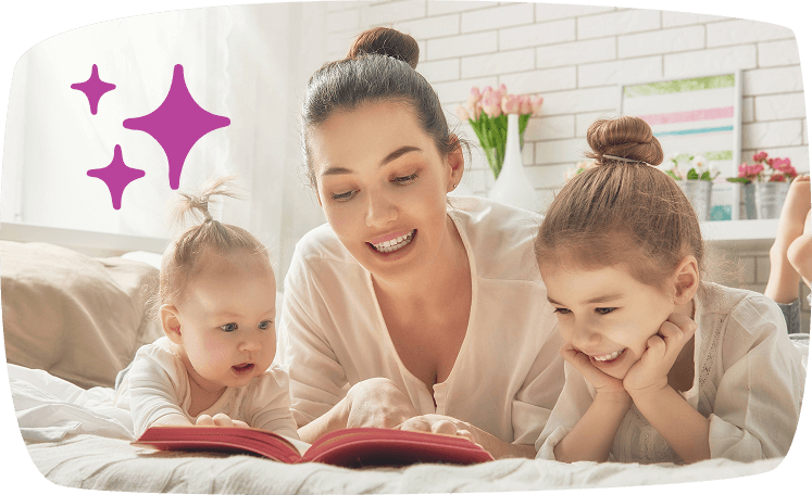 Madre leyendo un cuento en la cama junto a su bebé y su hija mayor, todos atentos y sonrientes.