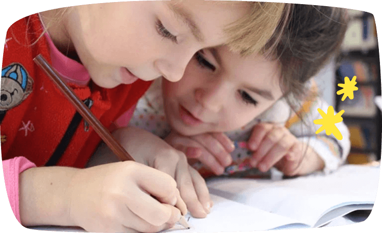 Dos niñas pequeñas escribiendo y dibujando en un cuaderno, concentradas y sonriendo mientras comparten la actividad.