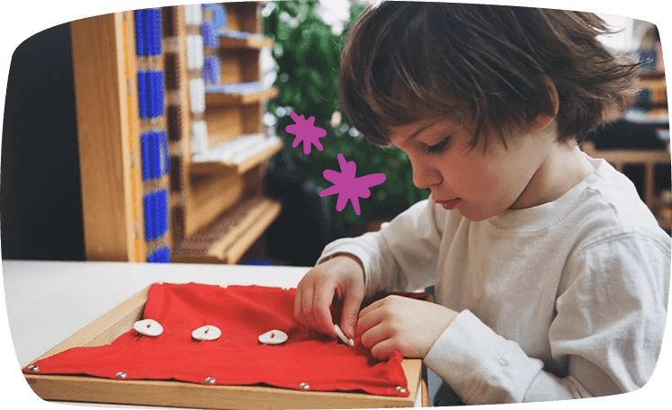 A child focused on buttoning a red Montessori dressing frame, practicing fine motor skills in a classroom setting.