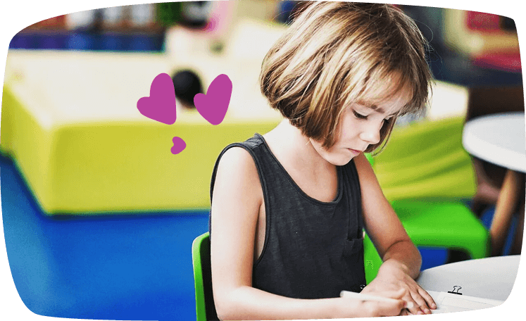 Niña concentrada escribiendo en un cuaderno en una sala de juegos, con decoración de corazones morados.