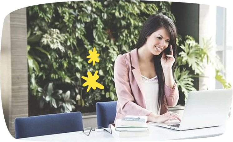 Mujer sonriendo mientras habla por teléfono frente a su portátil, rodeada de plantas.