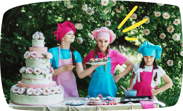 Niñas disfrazadas de reposteras presentando sus cupcakes en una mesa decorada al aire libre.