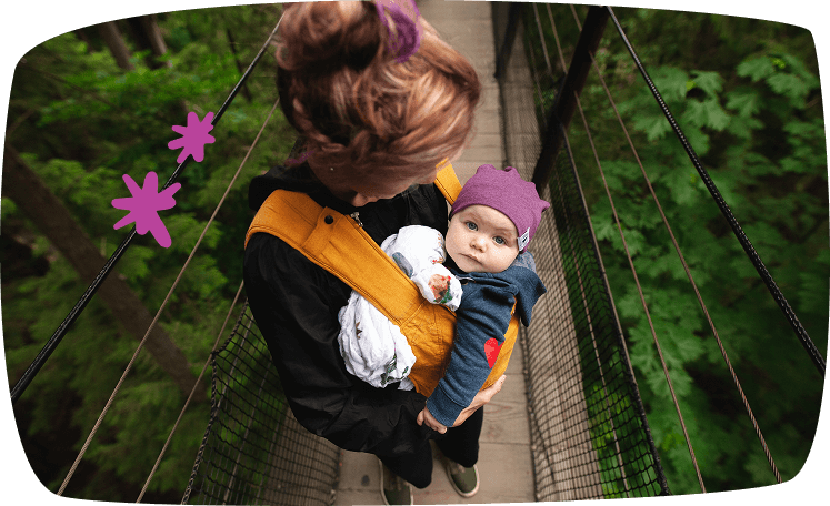 Madre sosteniendo a su bebé en una mochila ergonómica mientras cruzan un puente colgante en el bosque.