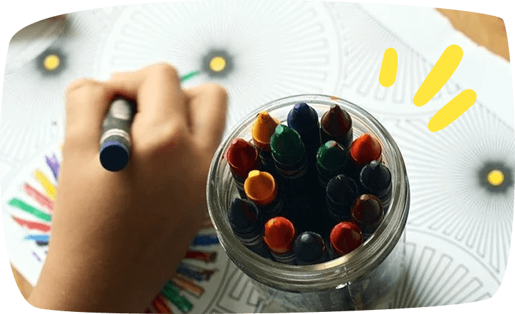 Niño dibujando con ceras de colores sobre un papel, con un bote lleno de ceras multicolores en primer plano. Actividad creativa para desarrollar la motricidad fina.