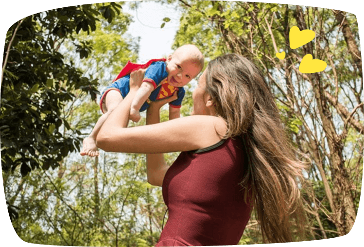 Mujer levantando en brazos a un bebé disfrazado de Superman al aire libre, rodeados de árboles, en un momento alegre y divertido.