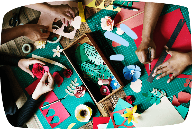 Manos de varias personas realizando manualidades con papel de colores, recortando y ensamblando flores sobre una mesa verde, con adornos gráficos de exclamación.