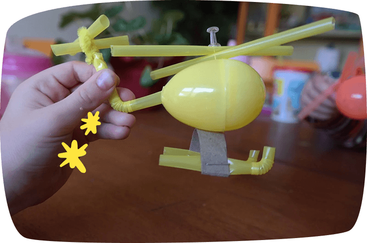 Helicóptero artesanal hecho con pajitas amarillas, un huevo sorpresa y limpiapipas, sostenido por una mano.