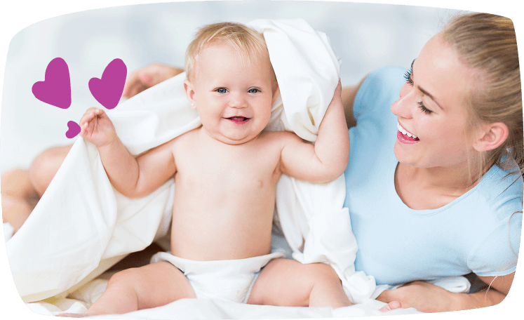 Madre sonriente juega con su bebé rubio, que se esconde bajo una manta blanca en la cama.